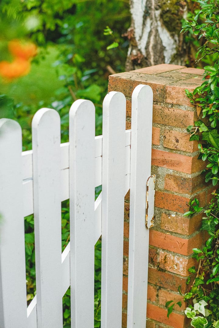 A serene garden entrance with a white wooden gate and red brick wall surrounded by lush greenery.